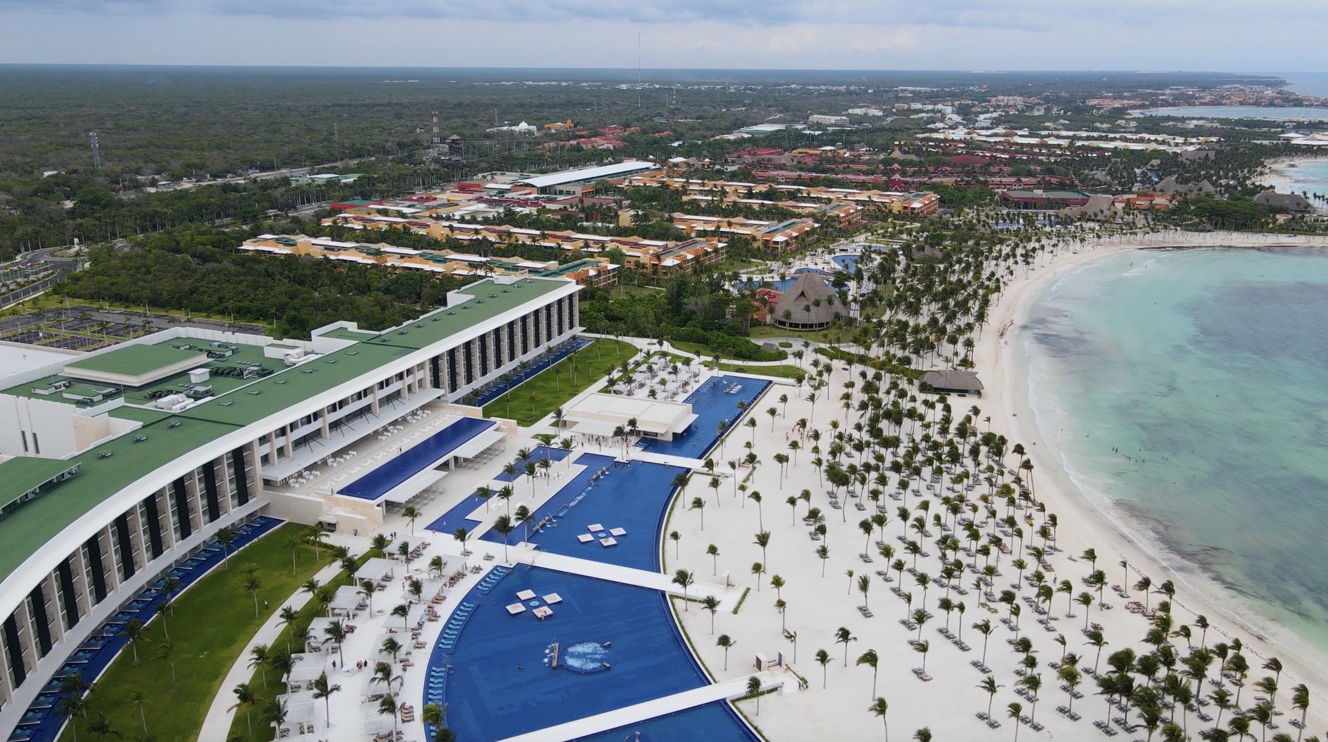 Aerial view of a beachfront Caribbean resort — turquoise pools, palm-lined promenade, and a white-sand beach meeting clear ocean