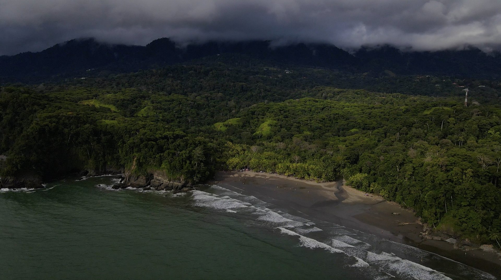 Aerial of a moody Pacific coastline in Costa Rica — dense jungle meeting a black-sand beach under a low cloud ceiling