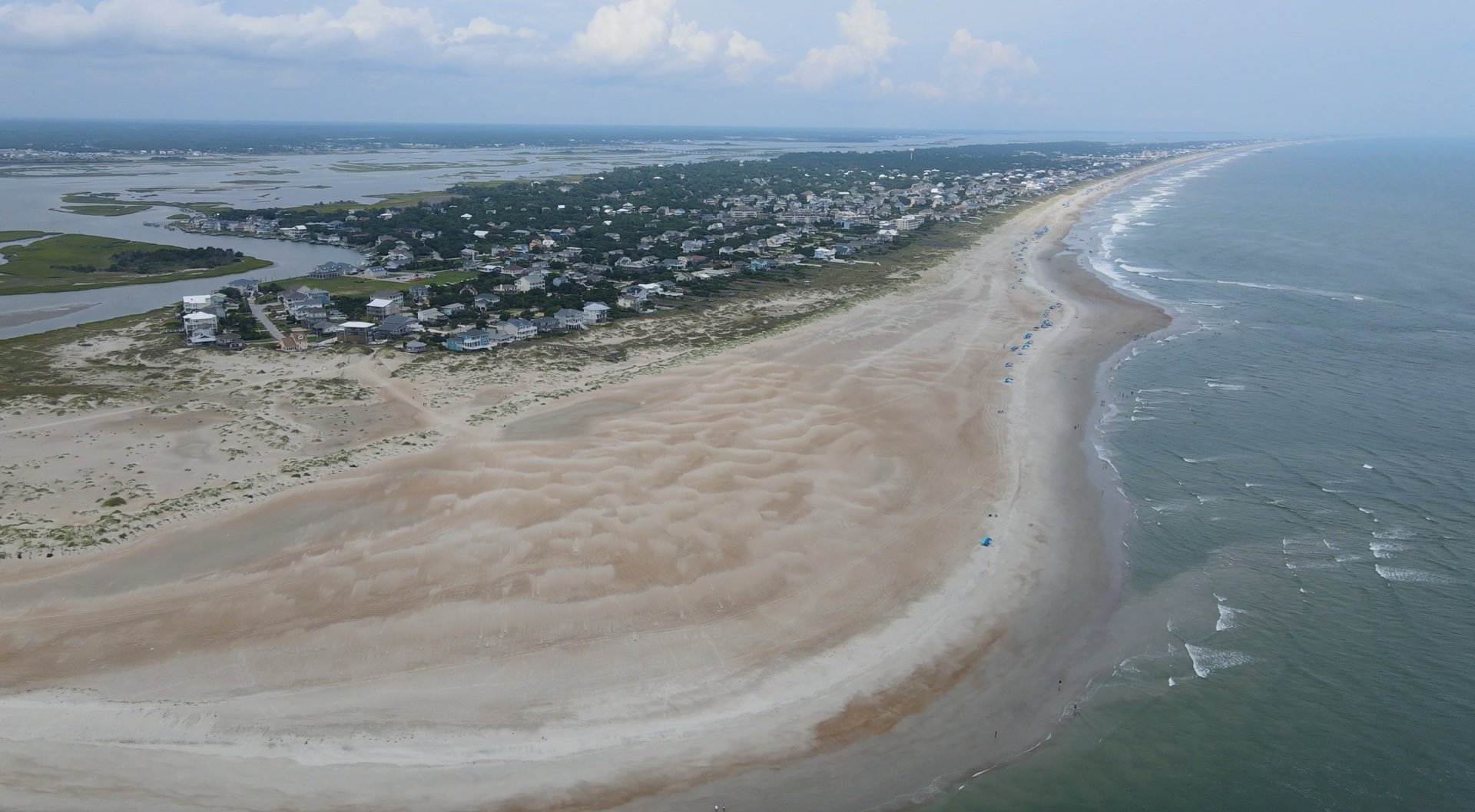 Aerial looking down a barrier island at the Outer Banks — sand stretching to the horizon with a narrow strip of beach town and inlet to one side
