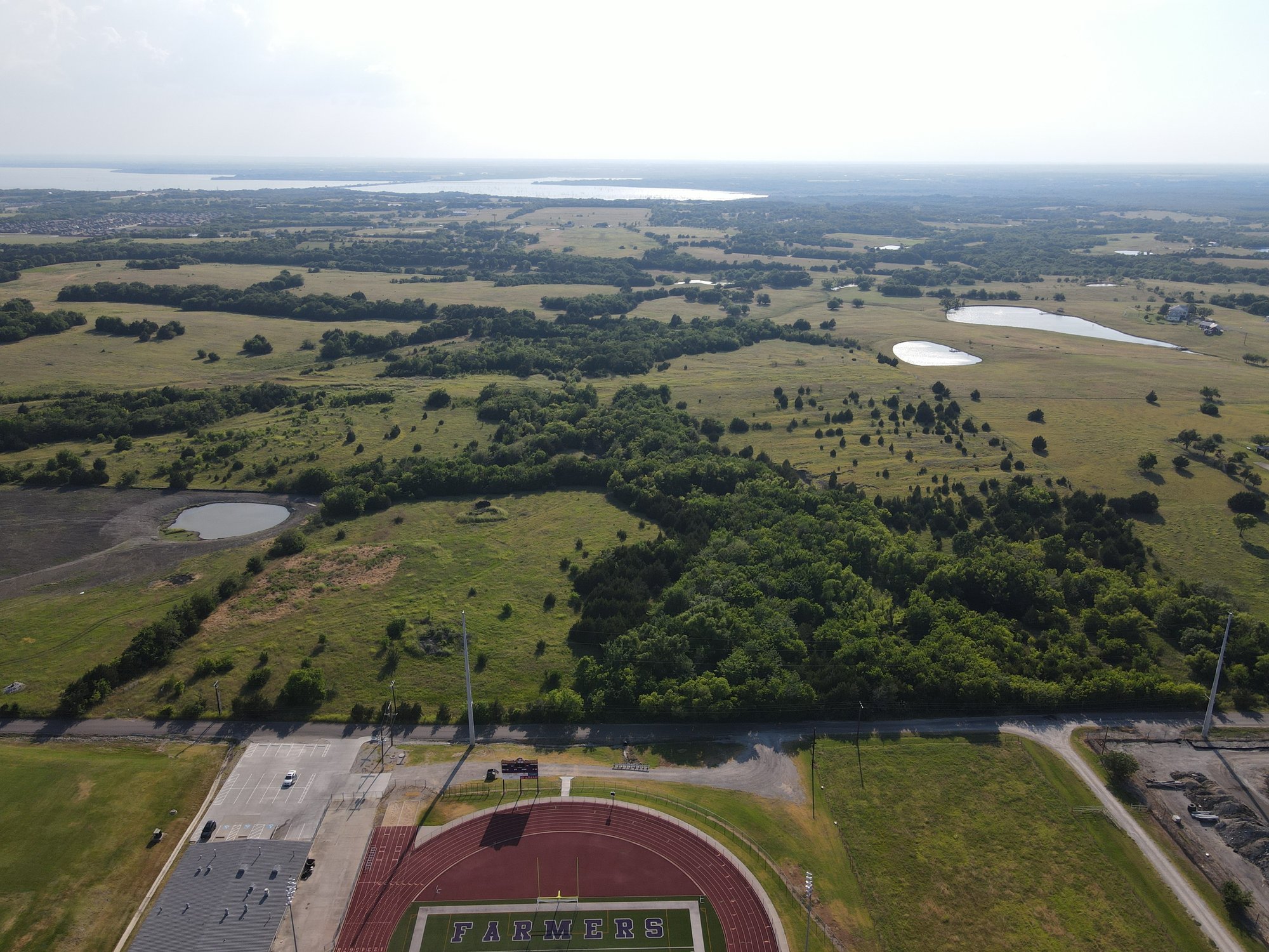 Mounger Creek aerial facing west — undeveloped tract adjacent to a school athletic complex, with rolling countryside and a distant lake
